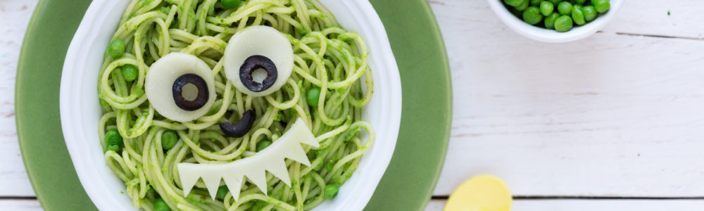 Fun food for kids - cute smiling face of green spaghetti monster served in a white bowl with eyes made of cheese and olives. Healthy vegetarian eating for children