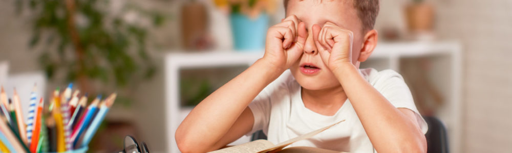 the child is tired of learning. home schooling, homework. the boy rubs his eyes from fatigue reading books and textbooks. a little boy student sitting at a table with books. vision problems